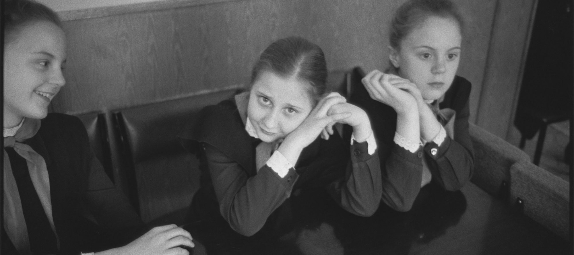 A black-and-white photograph of three light-skinned schoolgirls in dark, long-sleeved, matching uniforms. The girl on the right stares off, hands clasped by her cheek; the girl in the middle stares at the viewer with her head on her hands; and the girl on the left is laughing.