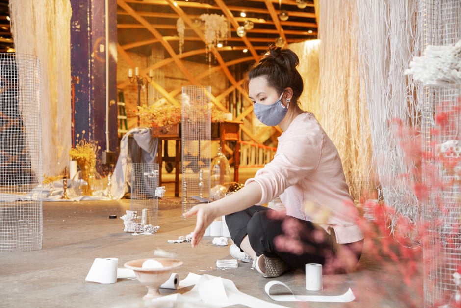 A light-skinned woman of Asian descent sits cross-legged on a concrete floor wearing a face mask, with a collection of detritus surrounding her, including rolls of toilet paper in different sizes, a clay bowl with an object in it, and others. Around and behind here textile works hang.