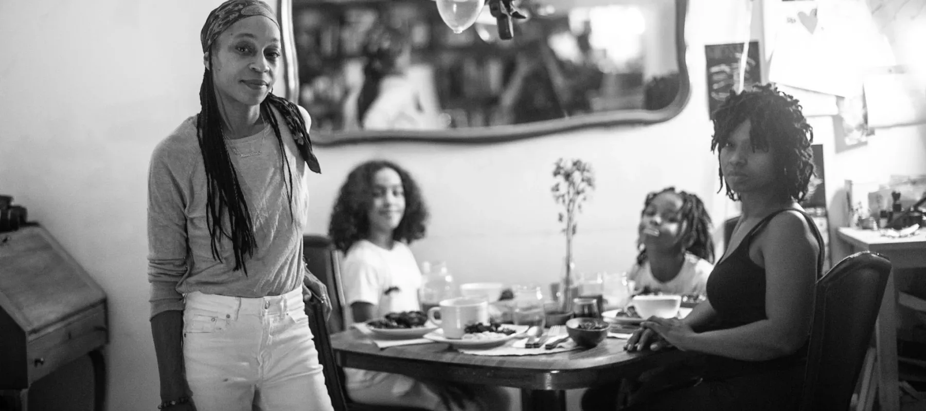 A black-and-white photograph of two women and two girls, all with medium-dark skin tones, together at a dining table set for dinner. A lamp hangs over the table and a mirror hangs on a wall behind them. The woman on the left is standing, while the other three sit at the table.