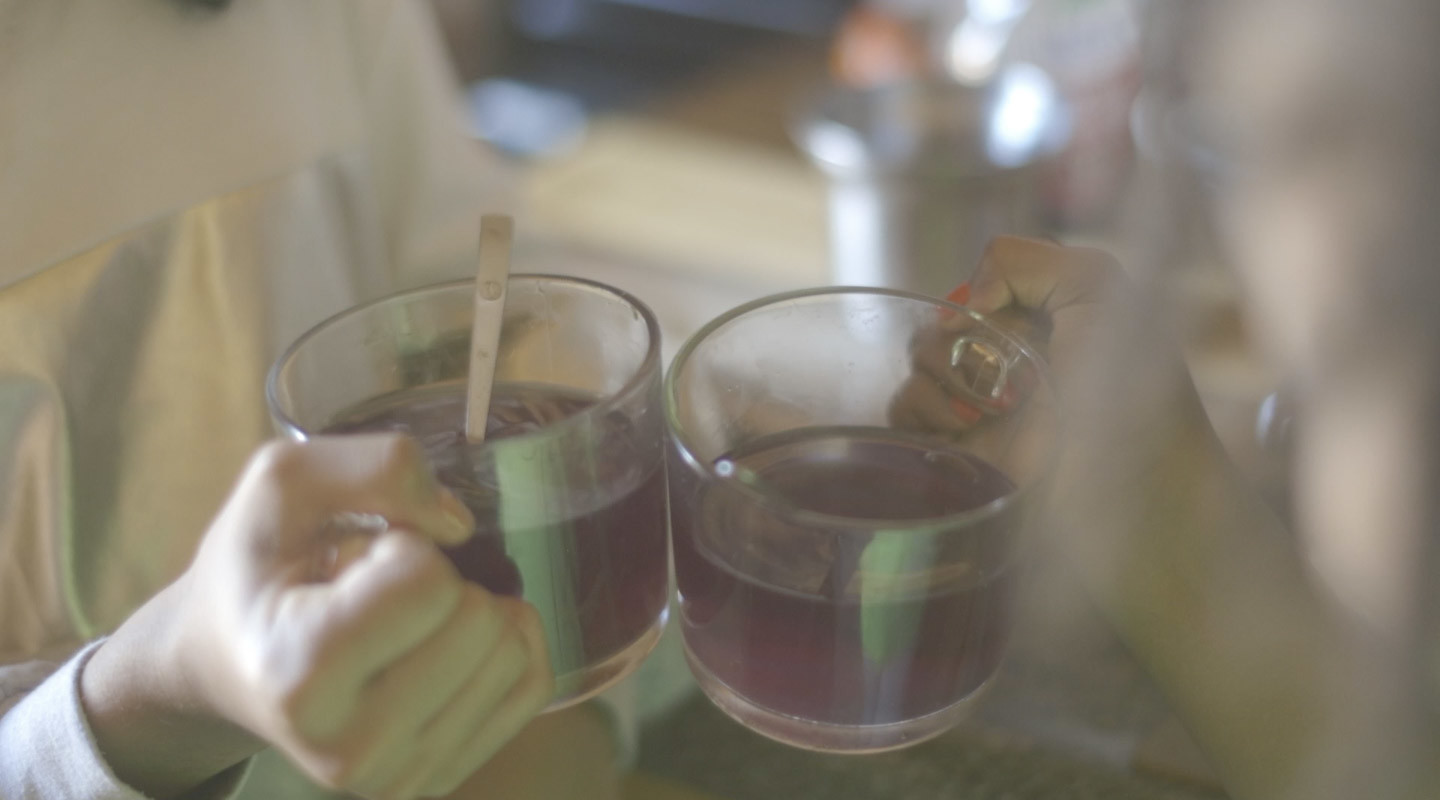 A photograph of two hands holding glass mugs of a purple tea. One has a small spoon in it.