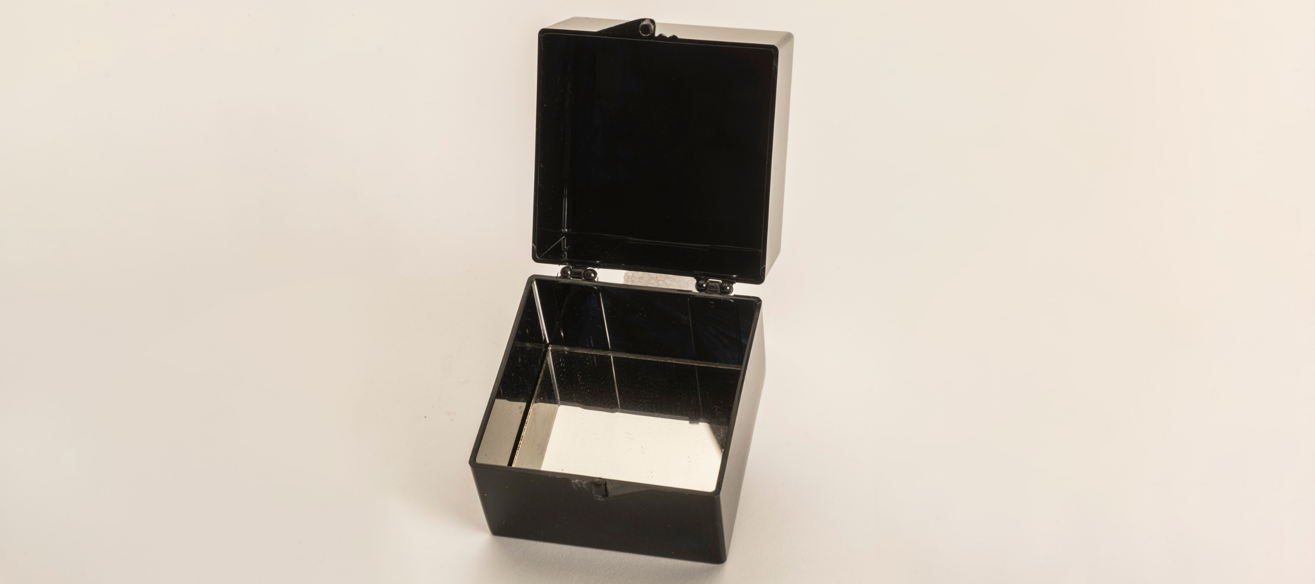 A square, black box open with its square, black lid facing up. It sits on a white background. Inside the box is a small mirror.