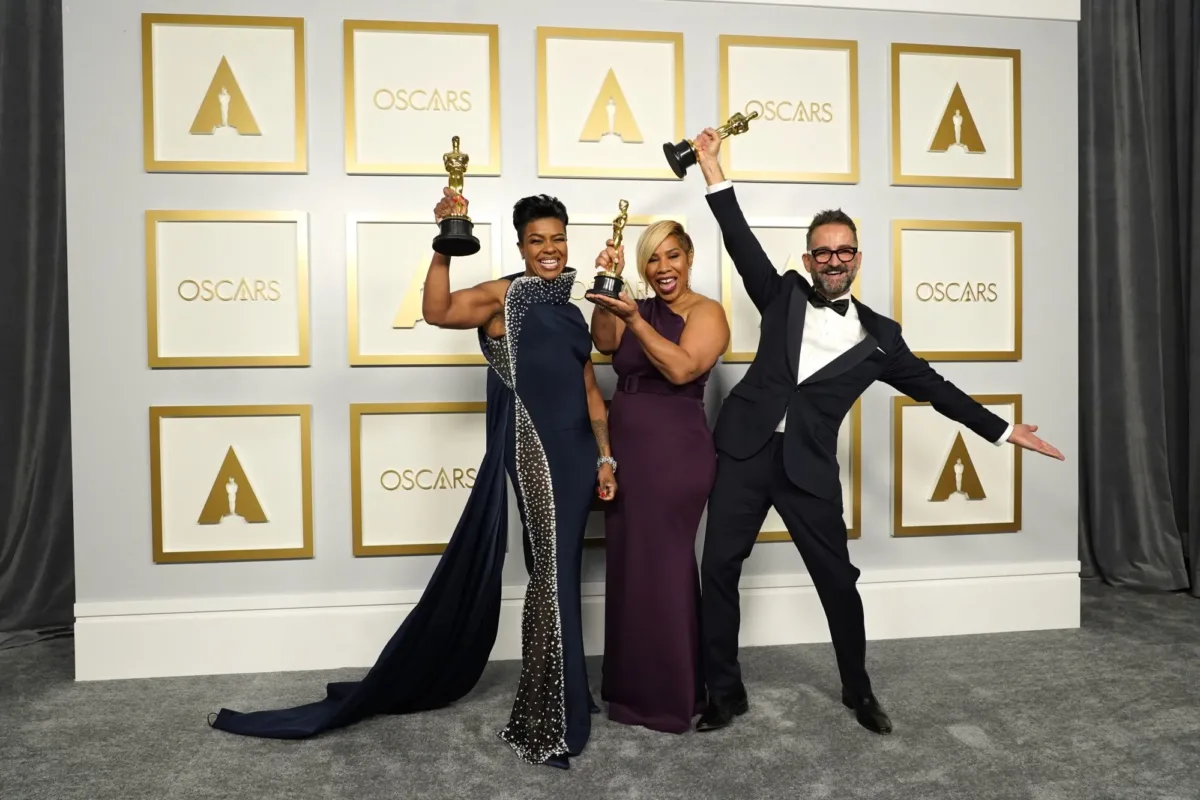 Two dark-skinned women and one medium skinned man celebrate in front of the Oscars photo backdrop. They are dressed in fancy evening wear and each hold up an Oscar statue with big smiles on their faces.