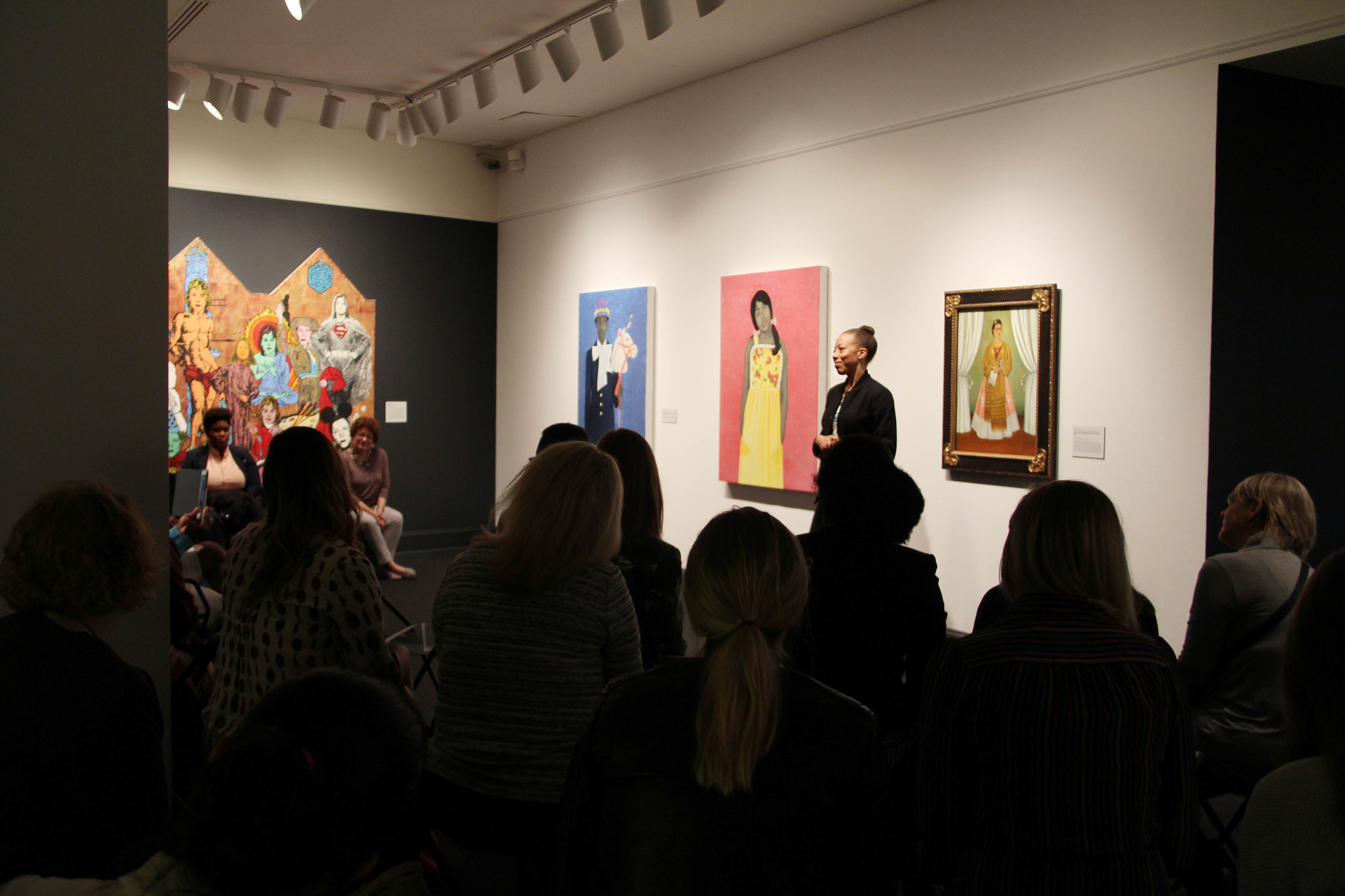 In a museum gallery, an artist with medium skin tone discusses her works to an audience.