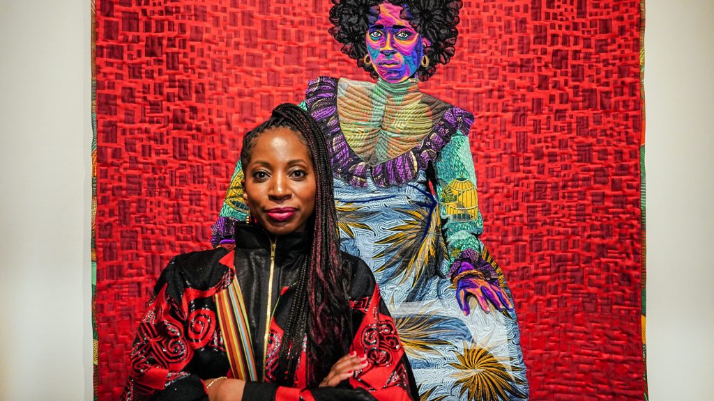 A dark-skinned woman with a high, braided ponytail stands in front of a colorful, fine art quilt depicting an image of a woman wearing a colorfully patterned dress and staring straight at the viewer. The real woman smiles slightly with her arms crossed across a vibrant red and black jacket zipped to her neck.