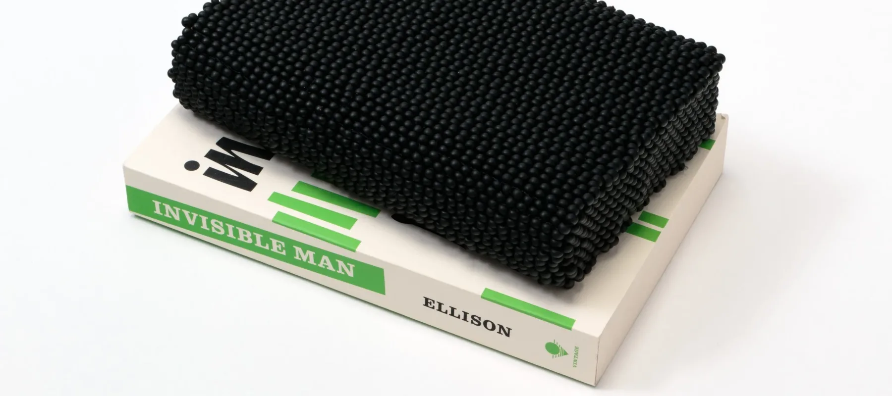 A paperback copy of Ralph Ellison's novel 'Invisible Man' sits on a white surface. The book features black text and short, green, vertical lines on a white background. Atop the book is a book-like shape comprising black beads.