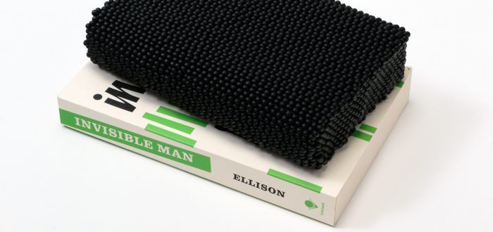 A paperback copy of Ralph Ellison's novel 'Invisible Man' sits on a white surface. The book features black text and short, green, vertical lines on a white background. Atop the book is a book-like shape comprising black beads.