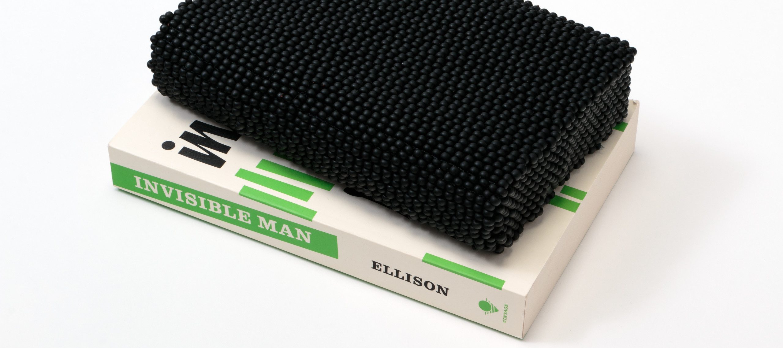 A paperback copy of Ralph Ellison's novel 'Invisible Man' sits on a white surface. The book features black text and short, green, vertical lines on a white background. Atop the book is a book-like shape comprising black beads.