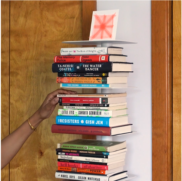 A tower of colorful hardback and paperback books on a white vertical bookshelf against wood and white walls.