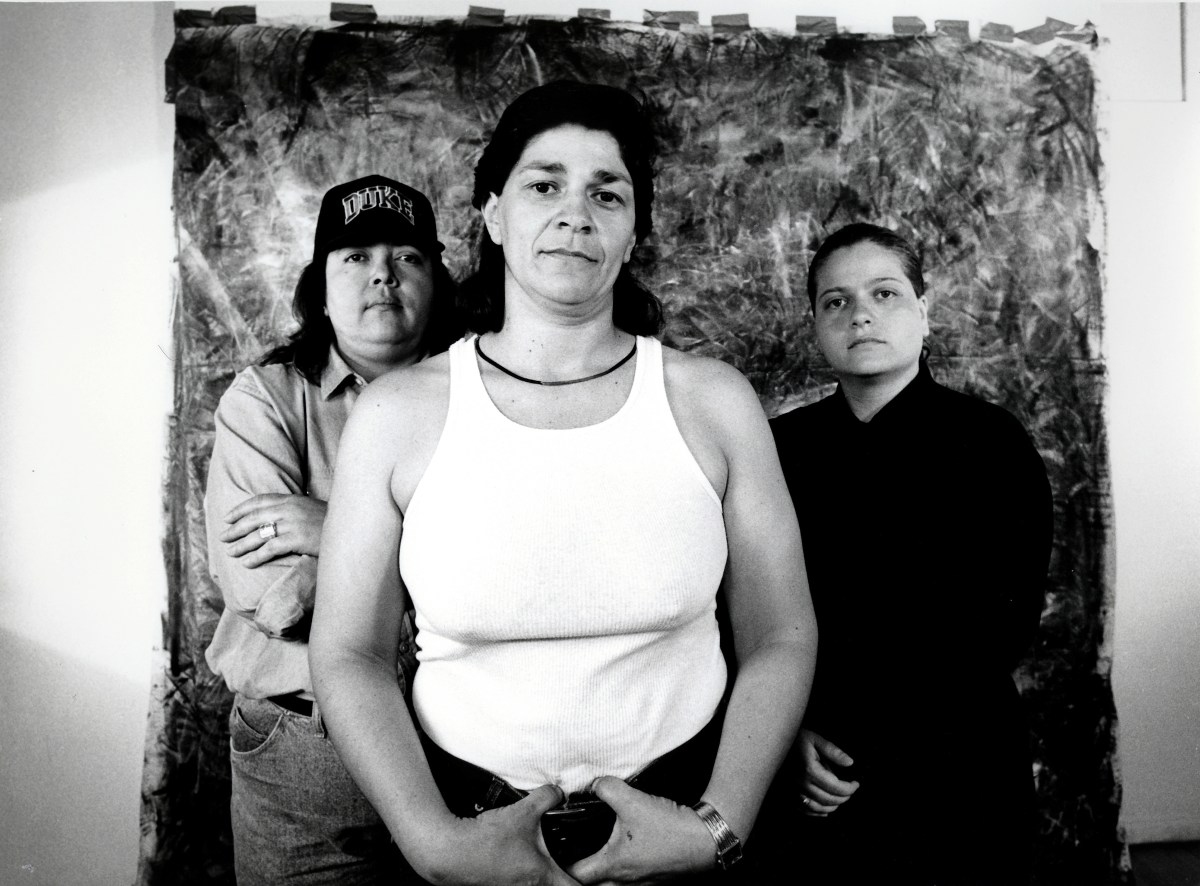 A black-and-white photograph of three Latino/a people standing in front of a taped backdrop. They look at the camera unsmiling and all wear casual clothing.