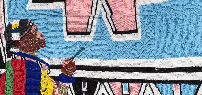 Tiny, colorful beads arranged in patterns to depict Esther Mahlangu painting a large artwork of geometric shapes in light pink and blue, black, white, yellow, and green. She is a dark-skinned adult woman wearing colorfully patterned robes.