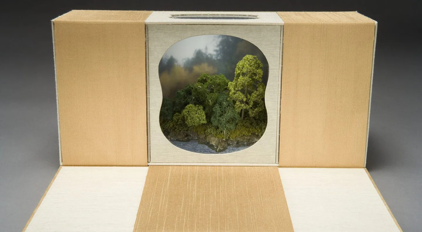 A beige and orange rectangular box sits with front flap open outward. A cutout window on the front side reveals a small landscape scene made of plastic model trees, rocks, and water.