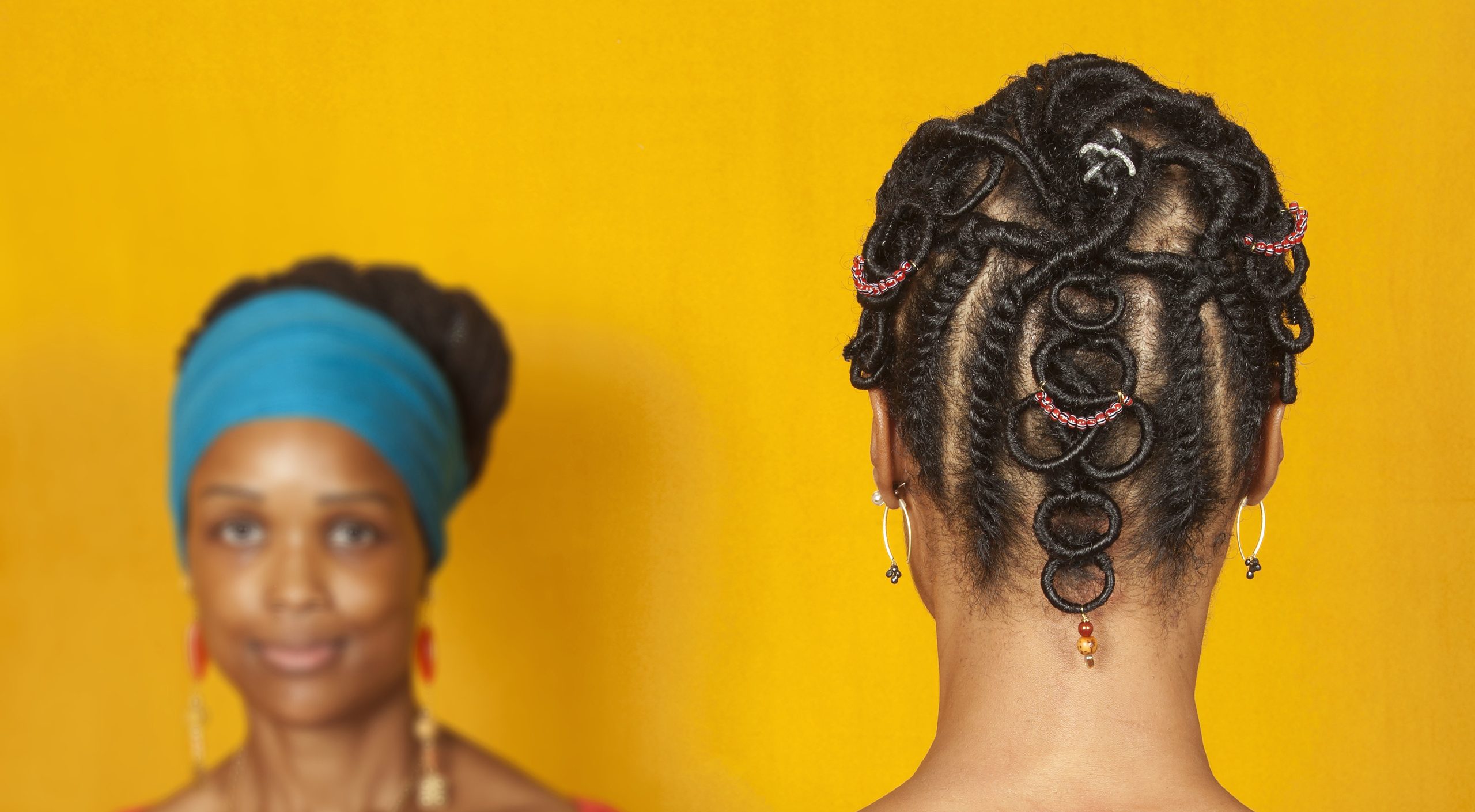 Two medium-dark skinned adult women stand in front of a goldenrod background. The woman on the left faces the viewer and wears a teal head wrap and a red top. The woman on the right faces away from the viewer, showcasing her dark hair braided into an elaborate pattern with colorful beads.