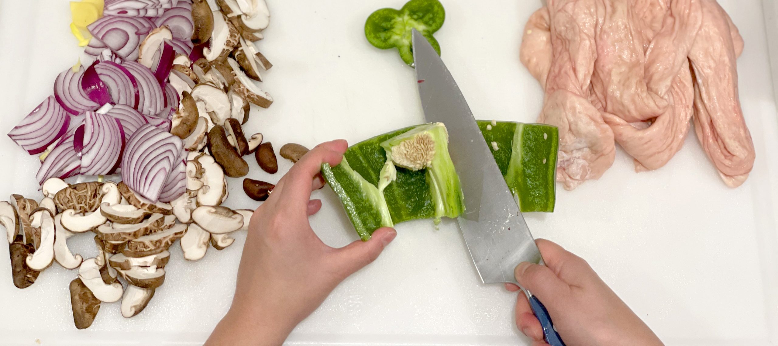 A pair of medium-light skinned hands slice the seeds out of a green pepper using a large knife on a white cutting board, seen from above. To the left is a pile of sliced mushrooms and red onions, and garlic. On the right is the raw skin of a chicken carcass.