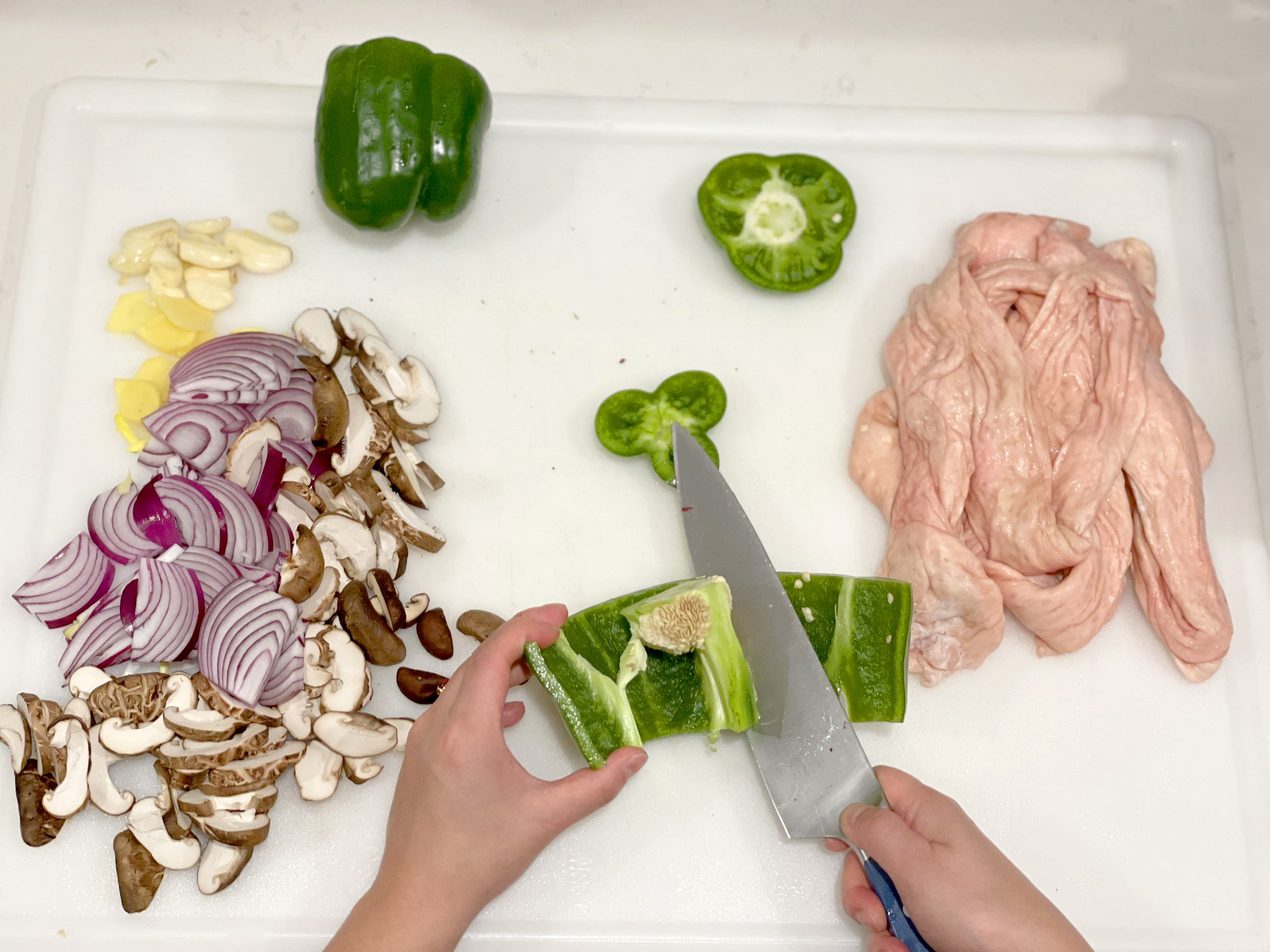 A pair of medium-light skinned hands slice the seeds out of a green pepper using a large knife on a white cutting board, seen from above. To the left is a pile of sliced mushrooms and red onions, and garlic. On the right is the raw skin of a chicken carcass.