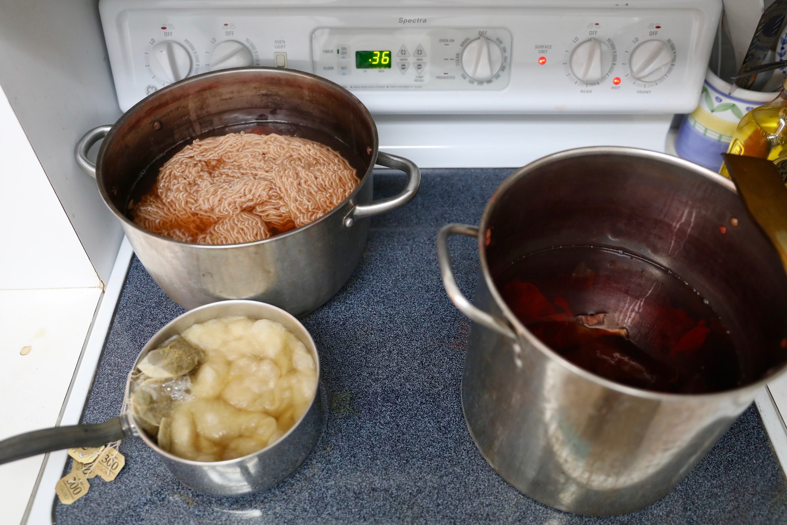 Atop an electric stove, three different sized stainless steel pots are filled with water and yarn or cotton. They are in the process of being dyed by tea bags, berries, and other organic matter.