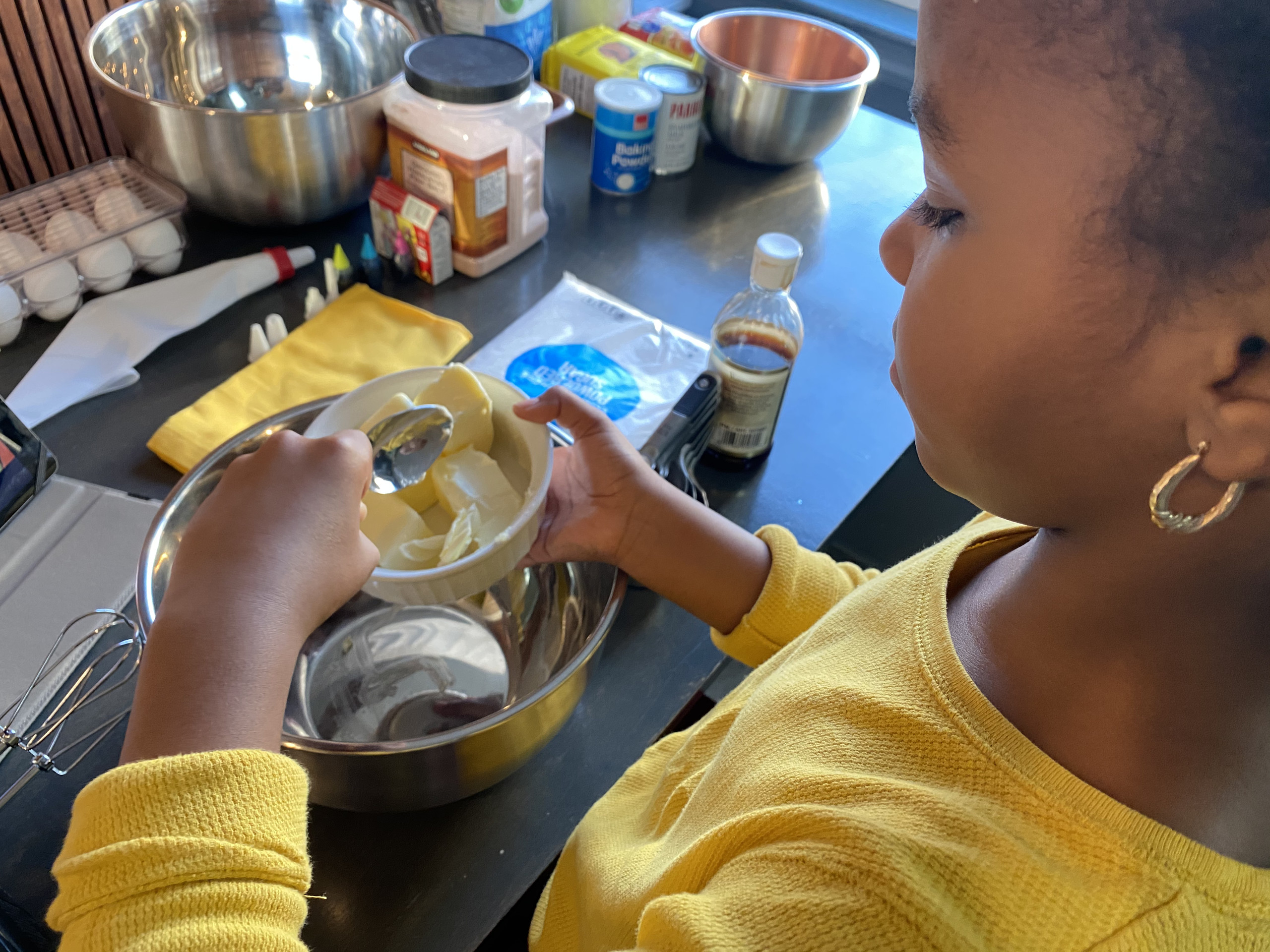 A view from over the shoulder of a medium-dark skinned girl baking at a counter that showcases a wide array of baking ingredients. She scoops chunks of butter into an empty, shiny, silver bowl, and wears earrings and a yellow shirt with sleeves rolled up.