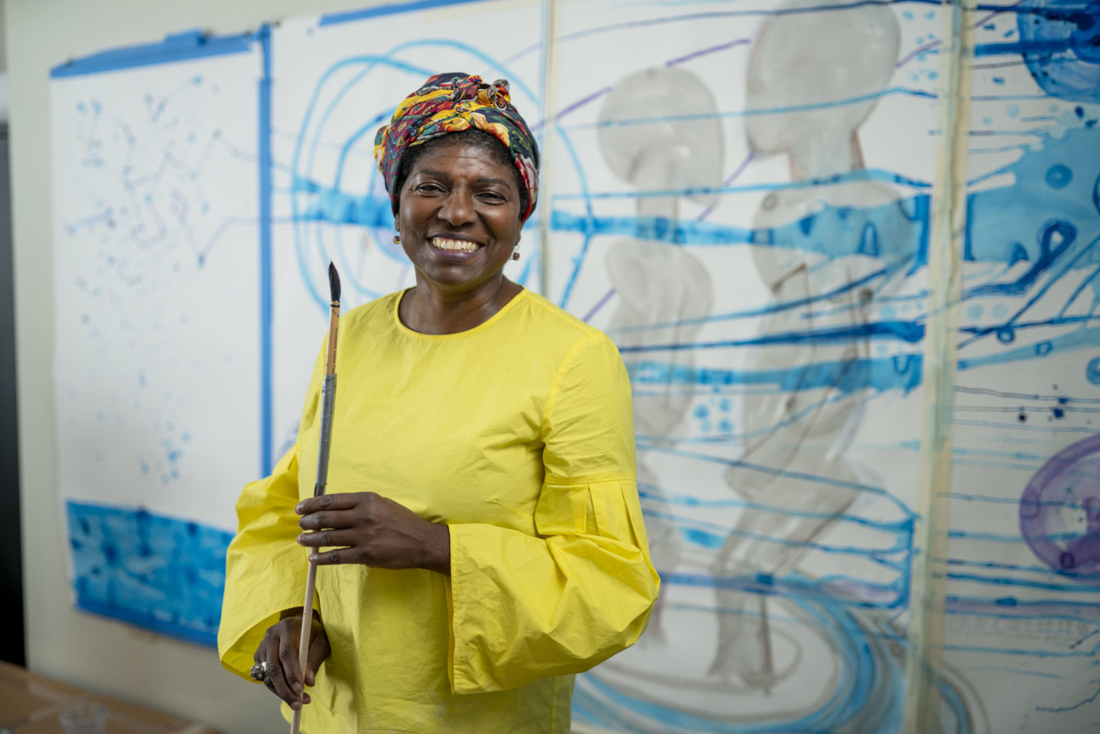 A dark-skinned woman in a bright yellow long barrel-sleeved dress stands in front of a wall of large artworks in process. She wears a colorful headwrap and smiles cheerfully while holding a long paintbrush.