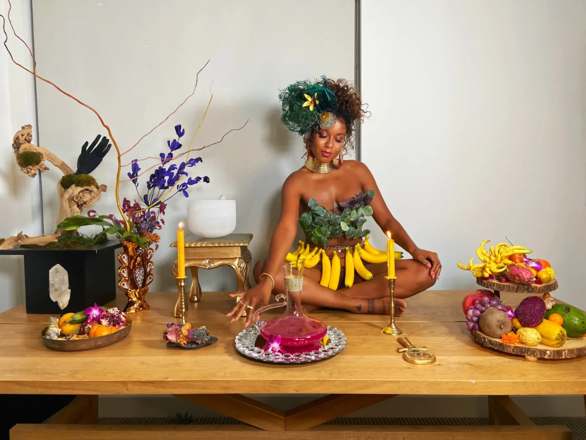 A medium-dark skinned adult woman sits cross-legged on a long wooden table set with platters of colorful fruit, candles, plants, and a pitcher of a pink liquid. She wears green leaves on her chest, gold jewelry, a skirt of bananas, and her hair pulled up with green feathers.