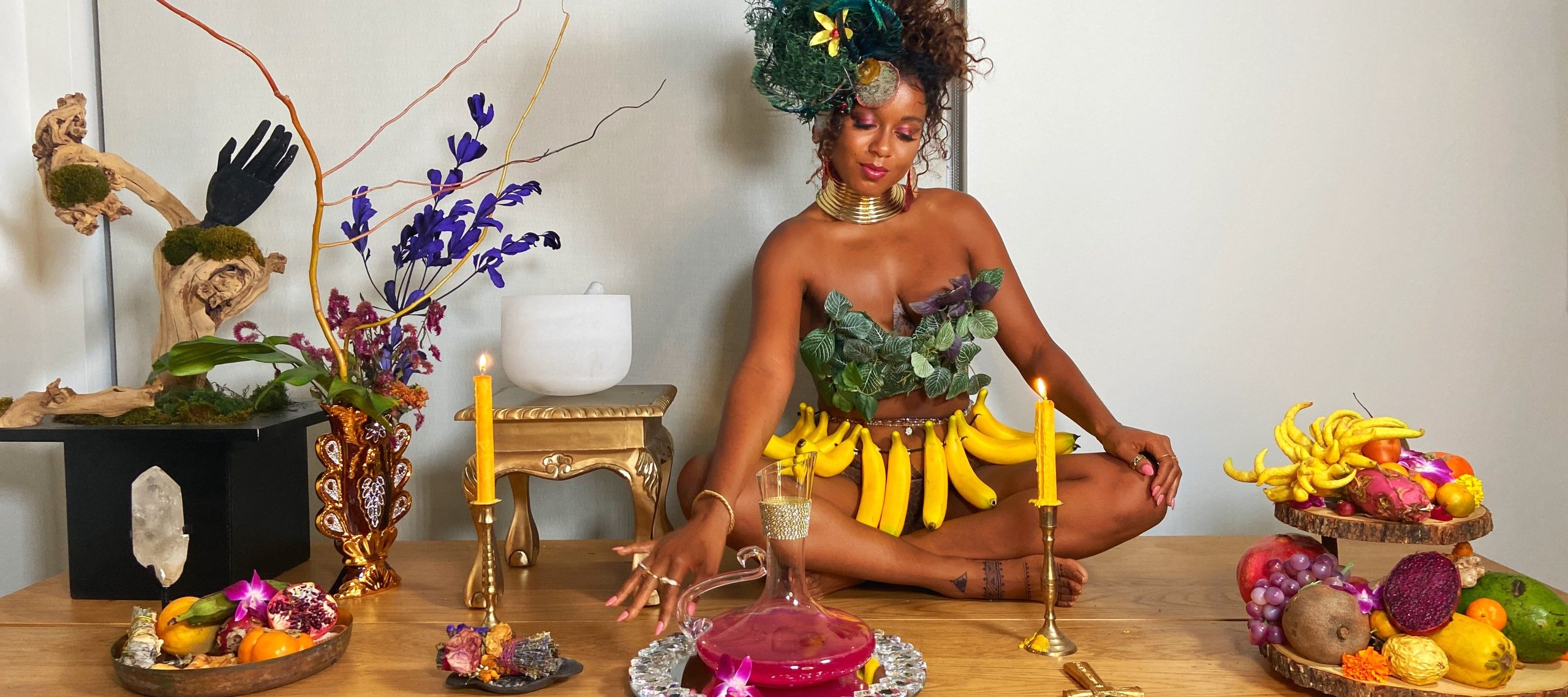 A medium-dark skinned adult woman sits cross-legged on a long wooden table set with platters of colorful fruit, candles, plants, and a pitcher of a pink liquid. She wears green leaves on her chest, gold jewelry, a skirt of bananas, and her hair pulled up with green feathers.