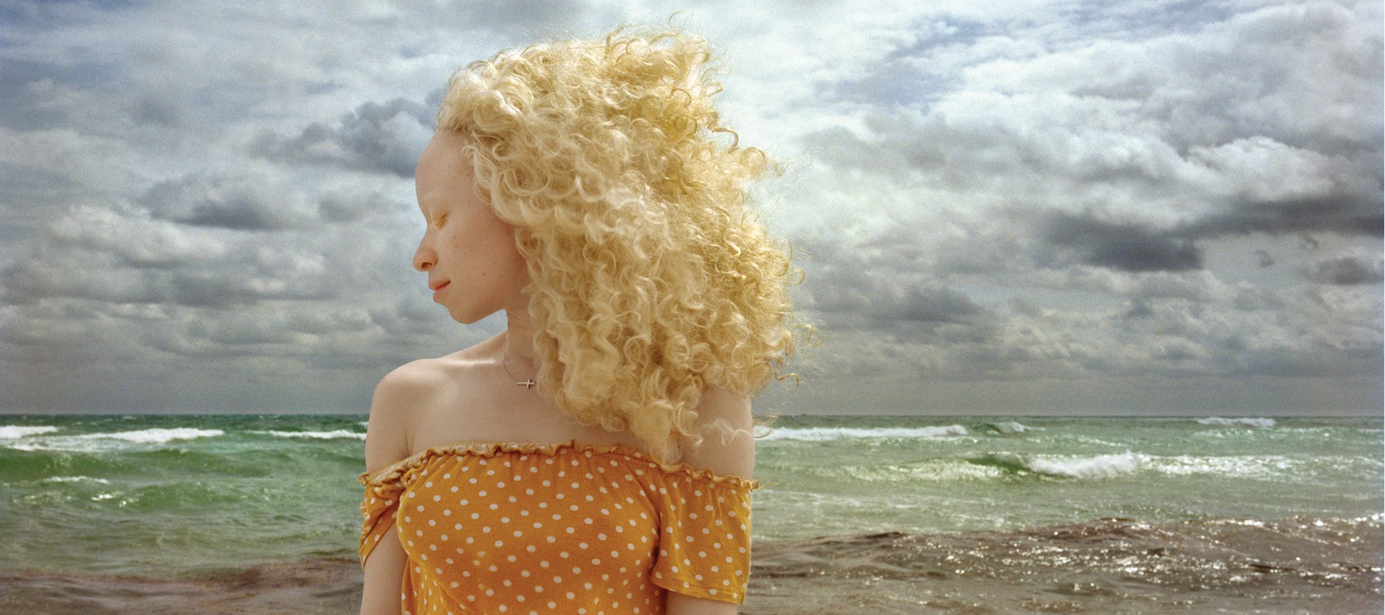In front of a stormy ocean, a woman with light skin and blonde, wind-blown hair stands with head turned in profile and eyes closed.