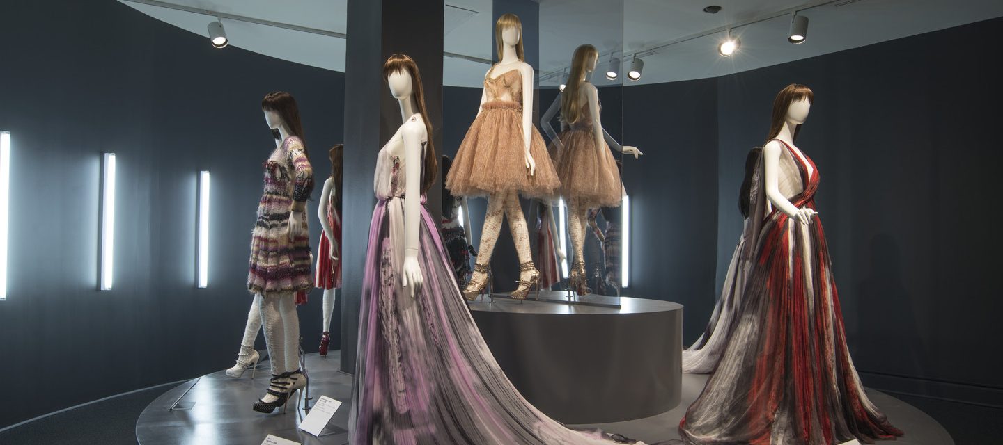 Five white mannequins in long formal dresses are standing in a dark space, illuminated by neon lights. The dresses are colorful and some have patterns on them.