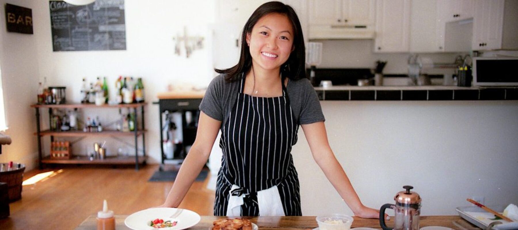 A light-skinned woman of Asian descent stands behind a large, wooden kitchen table that is full of two white dishes of finely plated food and various small, plastic to-go dishes., alongside a small bronze French Press coffee maker. The woman smiles and wears a grey tshirt under a black and white vertical striped apron.