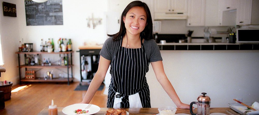 A light-skinned woman of Asian descent stands behind a large, wooden kitchen table that is full of two white dishes of finely plated food and various small, plastic to-go dishes., alongside a small bronze French Press coffee maker. The woman smiles and wears a grey tshirt under a black and white vertical striped apron.