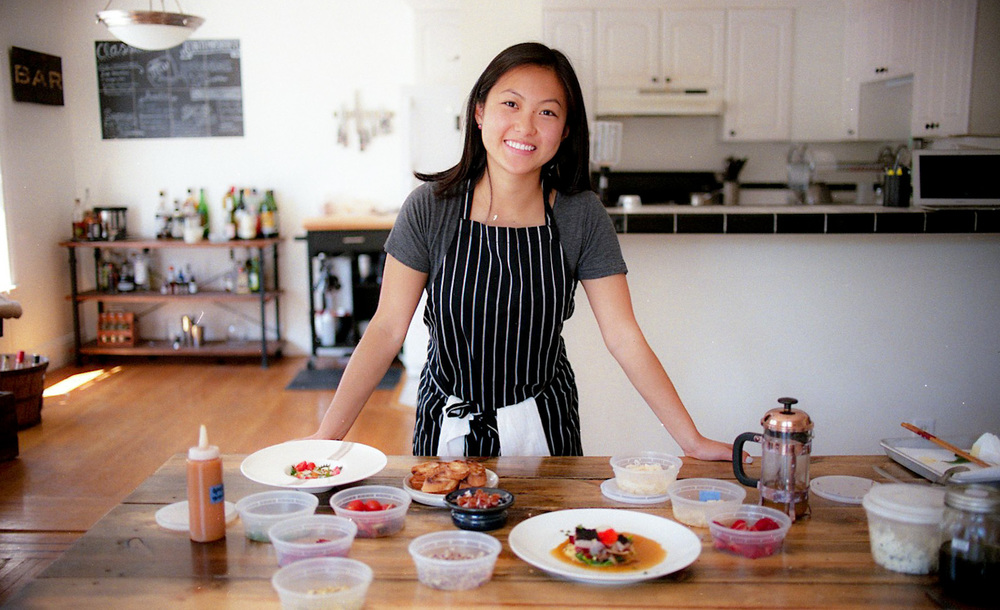 A light-skinned woman of Asian descent stands behind a large, wooden kitchen table that is full of two white dishes of finely plated food and various small, plastic to-go dishes., alongside a small bronze French Press coffee maker. The woman smiles and wears a grey tshirt under a black and white vertical striped apron.