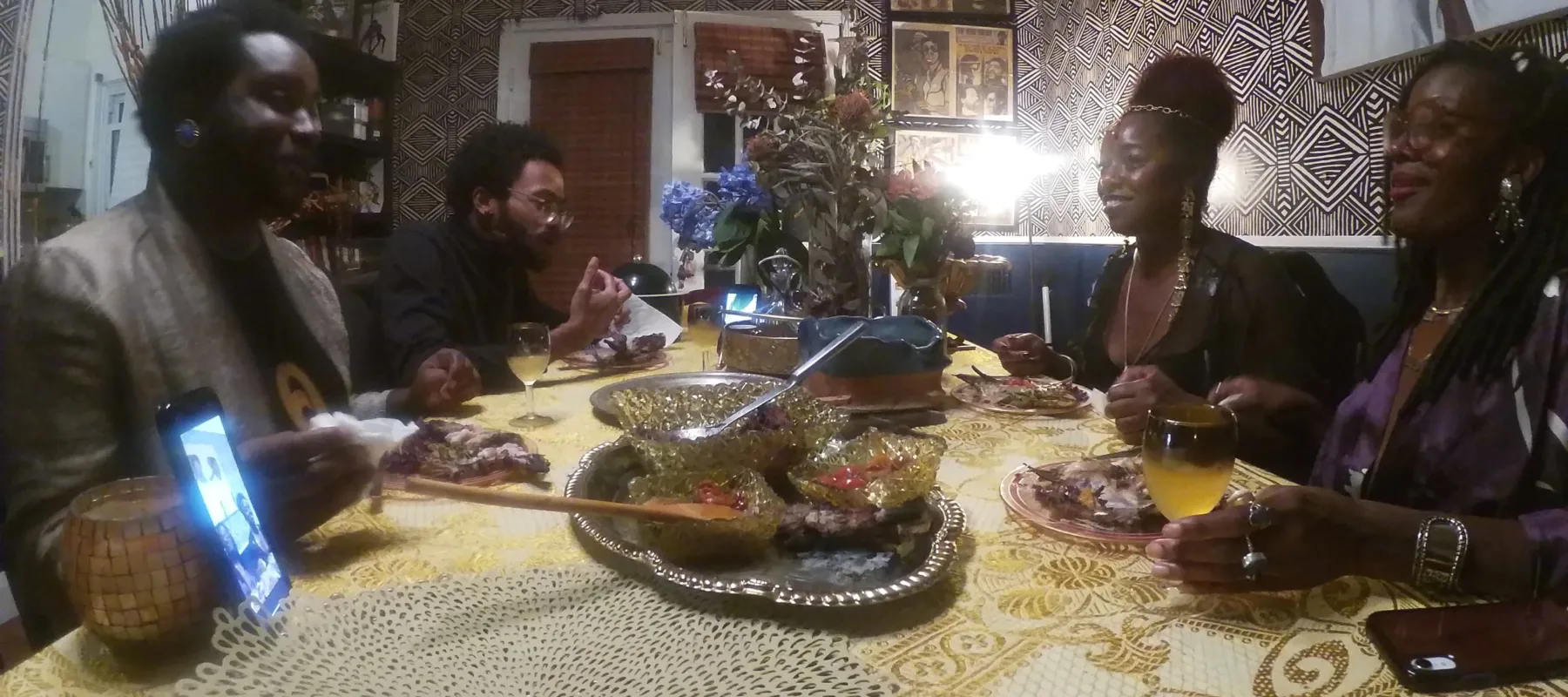 Four dark-skinned adults dressed up in suit jackets, nice tops, and jewelry sit around a table set with a yellow patterned tablecloth and meal, two on each side. A phone stands on the table, displaying other people calling in. The walls are covered in art and patterned wallpaper.