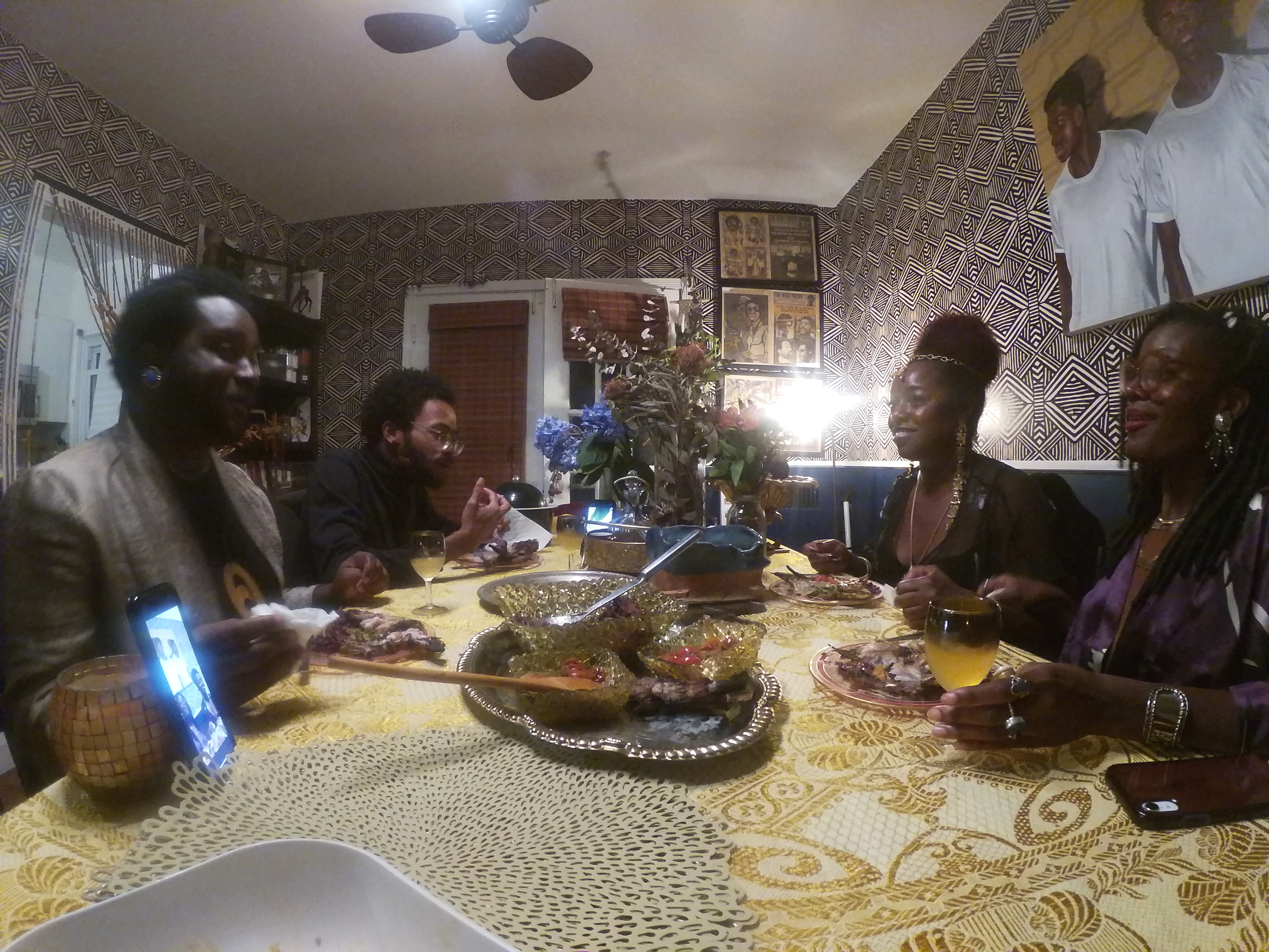 Four dark-skinned adults dressed up in suit jackets, nice tops, and jewelry sit around a table set with a yellow patterned tablecloth and meal, two on each side. A phone stands on the table, displaying other people calling in. The walls are covered in art and patterned wallpaper.