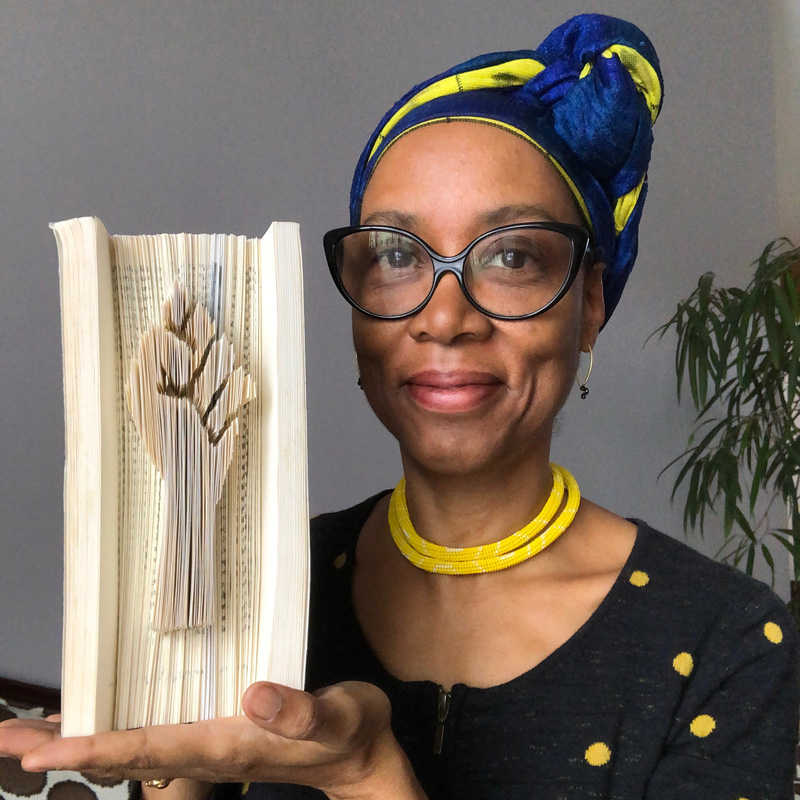 A medium-skinned woman wearing a blue and yellow head wrap smiles at the camera and holds up a book vertically. In the pages a solidarity first has been carved.