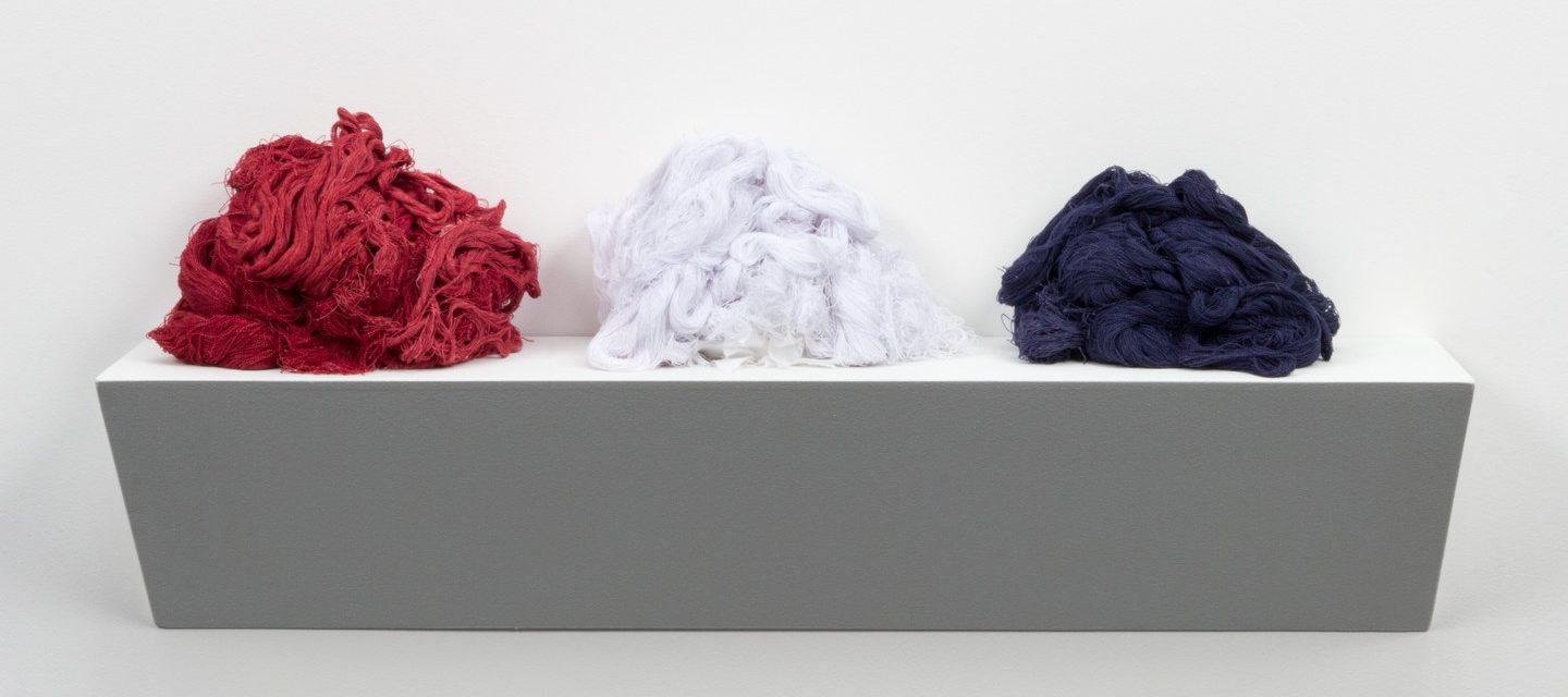 Three piles of thread from an unravelled American Confederate Battle Flag on a white base resembling a shelf. The thread piles are red, white, and blue.