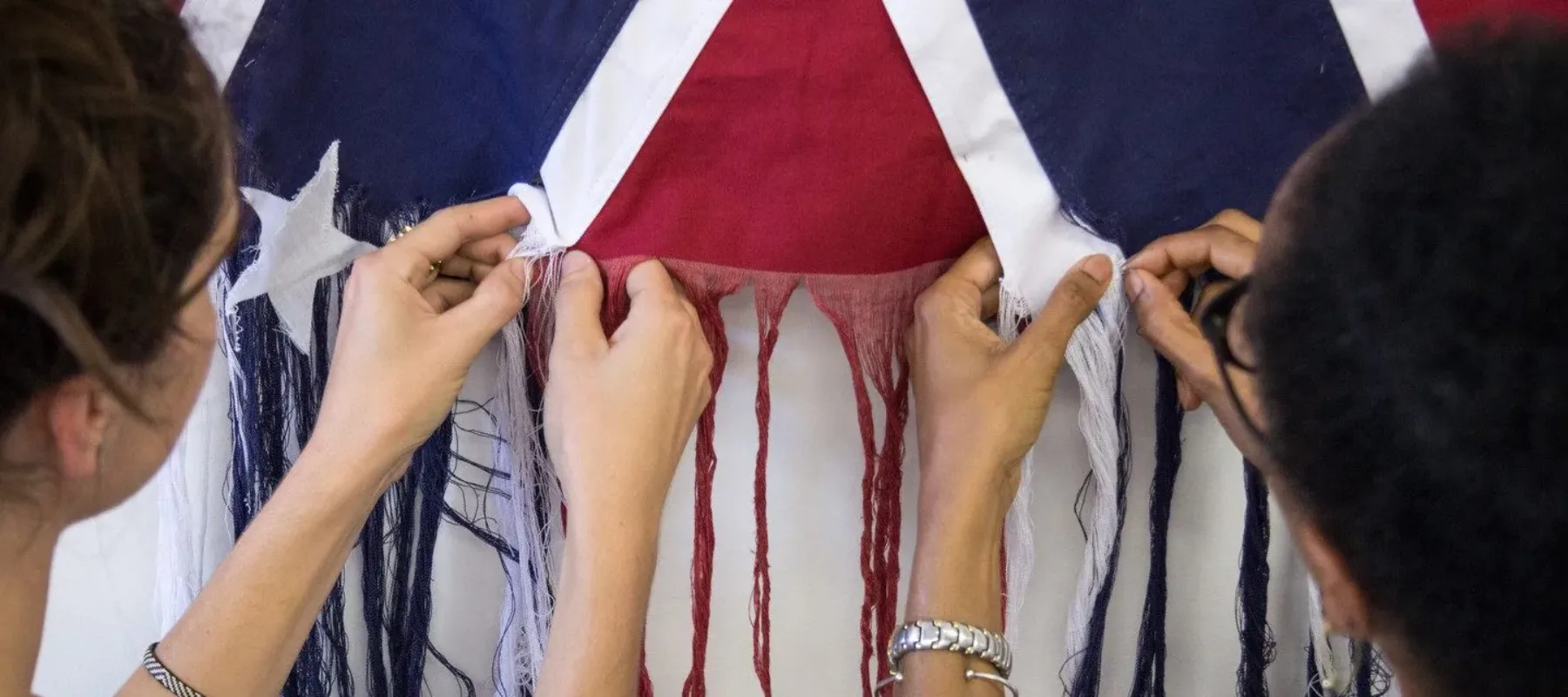 A photograph of two figures standing side by side, unraveling the threads of an American Confederate battle flag. The figure on the left has light skin, and the figure on the right has darker skin. They face away from the camera, with their hands in the center of the image pulling loose the threads.