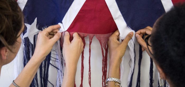 A photograph of two figures standing side by side, unraveling the threads of an American Confederate battle flag. The figure on the left has light skin, and the figure on the right has darker skin. They face away from the camera, with their hands in the center of the image pulling loose the threads.