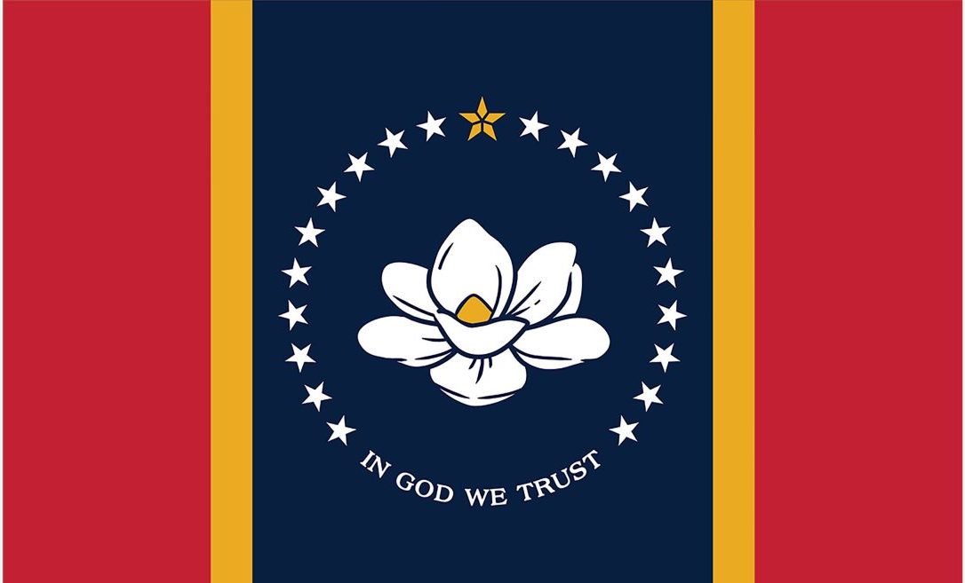 A state flag features a central, white magnolia with a gold center, surrounded by 20 white stars and a gold five point star at the top. Below the magnolia "In God We Trust" is written in all caps. The stars and magnolia are atop a navy blue background, which is framed by narrow gold stripes on either side, followed by rectangular red stripes on either side of that.