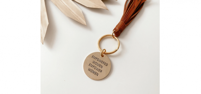 A small, circular brass keychain pendant is engraved with the phrase "EMPOWERED WOMEN EMPOWER WOMEN" in all caps. It hands on the end of a leather tassel.