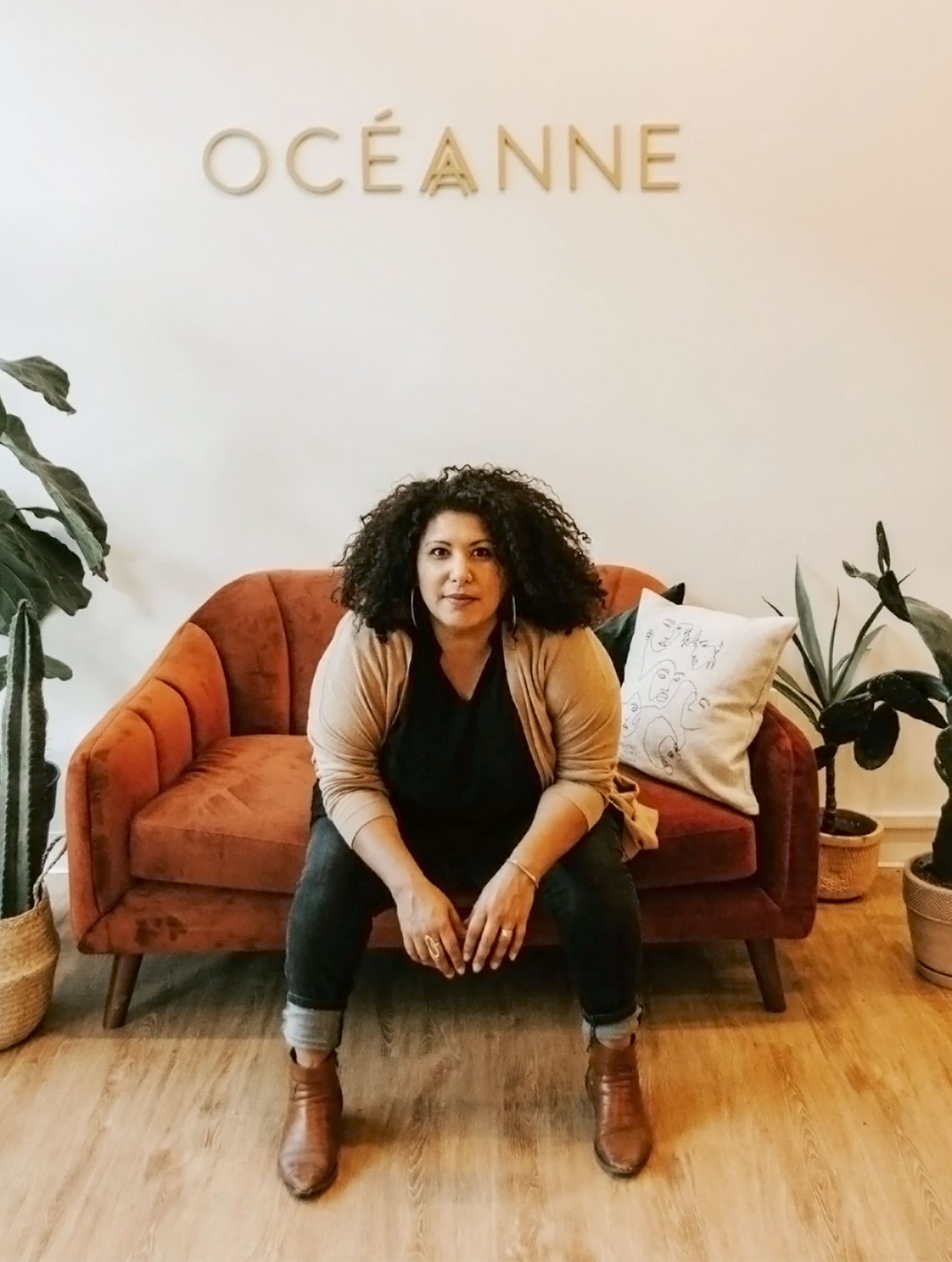 A medium-skinned woman with curly, shoulder-length black hair sits on a burnt orange couch, leaning forward with her forearms on her knees. She stares straight at the camera.