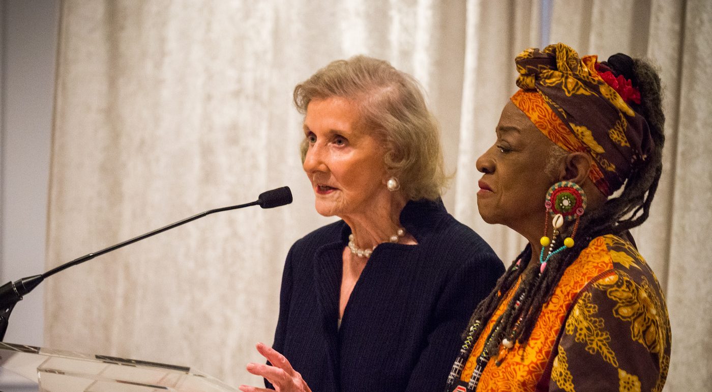 A light-skinned older woman speaks into a black microphone at a podium. Standing next to her is a black woman wearing a colorfully patterned matching headwrap and shirt and large beaded earrings.