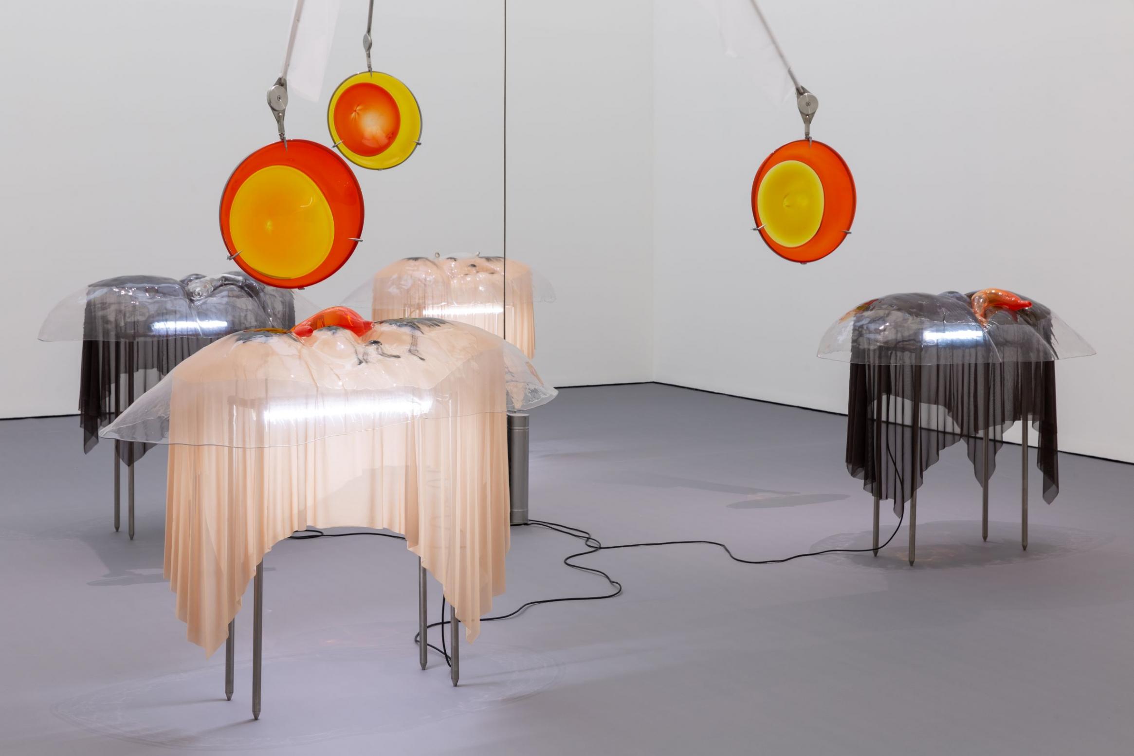 An installation photograph of abstract sculptures in a sparse gallery. Tables seem to be covered with pink and black latex fabric; circular orange and yellow orbs hang from the ceiling. Black cord is laid between the tables.