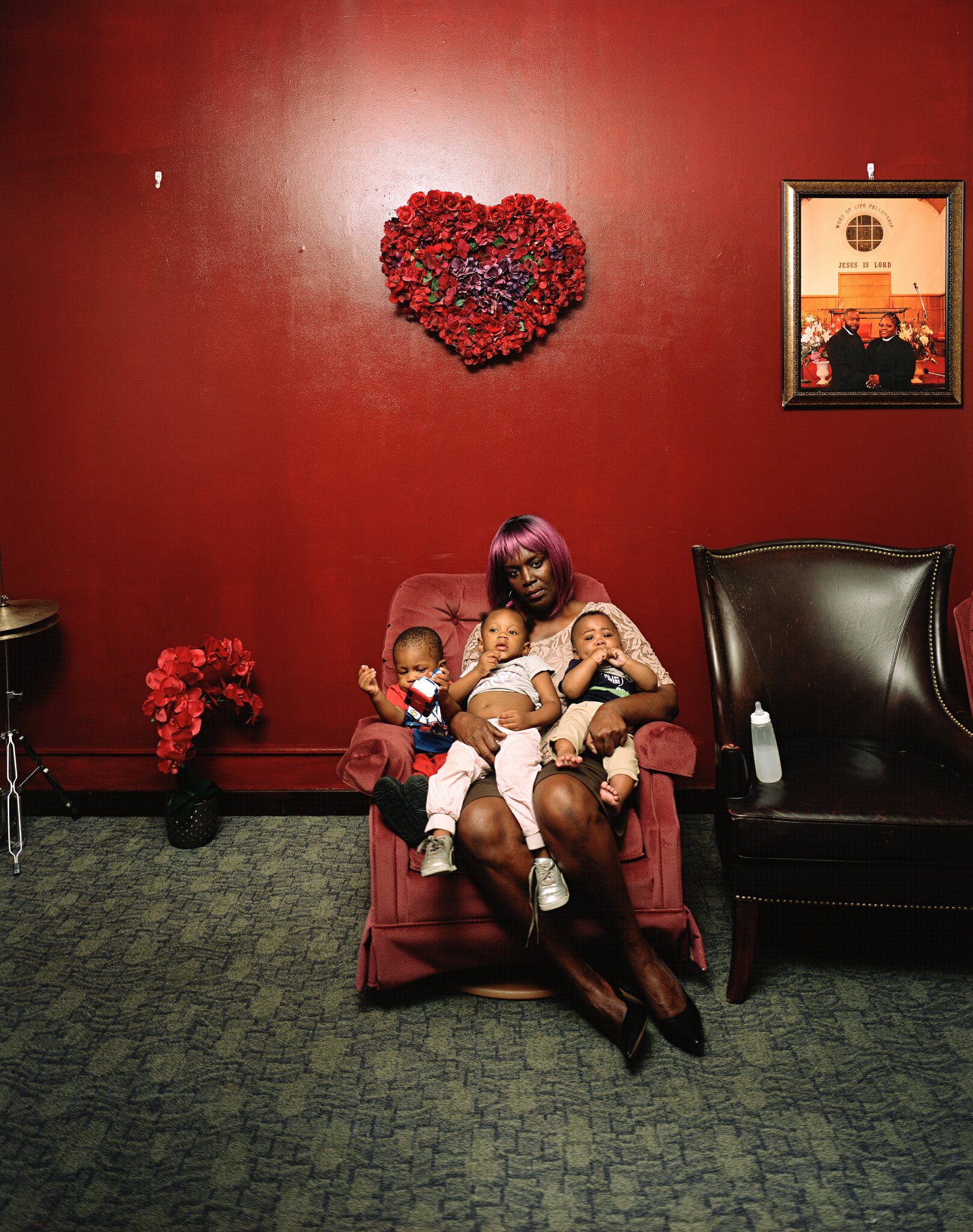 A dark-skinned woman sits on a velvet, mauve recliner chair holding three medium-skinned babies in her lap, all around the ages of 1 to 2. The wall behind her is painted a dark, shiny red, and above her a heart made from flowers hangs as a centerpiece.