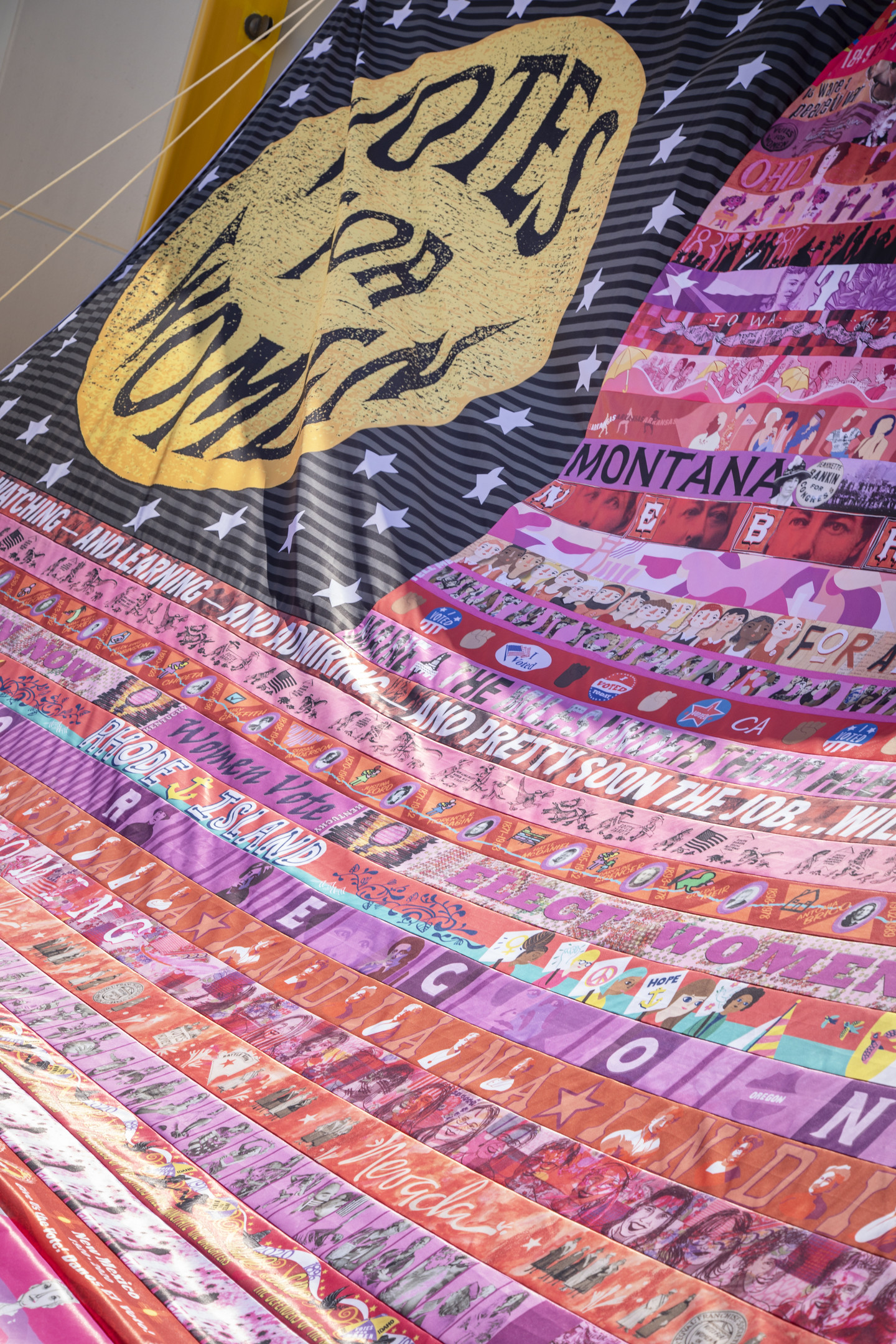 Detail of a flag that resembles an American flag with a gold oval sitting on a black rectangular background in the upper left corner of the flag. The gold oval contains the words "Votes for Women" in black text, the black background is filled with white stars. The stripes of the flag are made up of multicolored strips of fabric containing campaign slogans, images, and the names of several US states including Wisconsin, Kansas, New York, Ohio, Texas, Iowa, Montana, Nebraska, Maine, Rhode Island, Oregon, Indiana, Wyoming, Nevada, New Mexico, Oklahoma, and West Virginia.