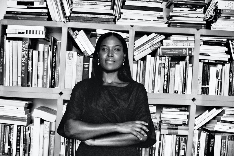 A high-contract black-and-white photo of a dark-skinned woman standing with her arms folded across her chest in front of a wall of cube book cases filled to the brim with books.
