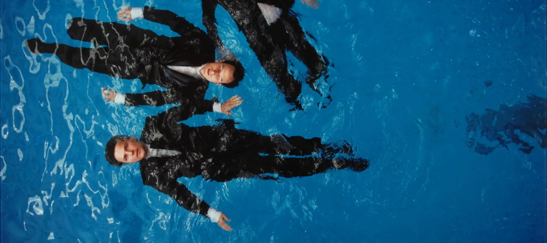 A photograph of four light-skinned figures with short brown hair, wearing black suits with white shirts, floating on their backs in a deep blue pool. The viewer looks down on the figures from above. One figure has their eyes closed, the rest have their eyes open.