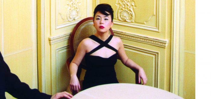 A light-skinned woman of Asian descent is dressed up in a fancy black dress, sitting at a table in a room with ornate yellow walls. Her hair is in an updo. She stares unsmiling at the camera, to her left is a male companion in a suit, thought he has been cut from the frame and all that shows his is arm and half is body.