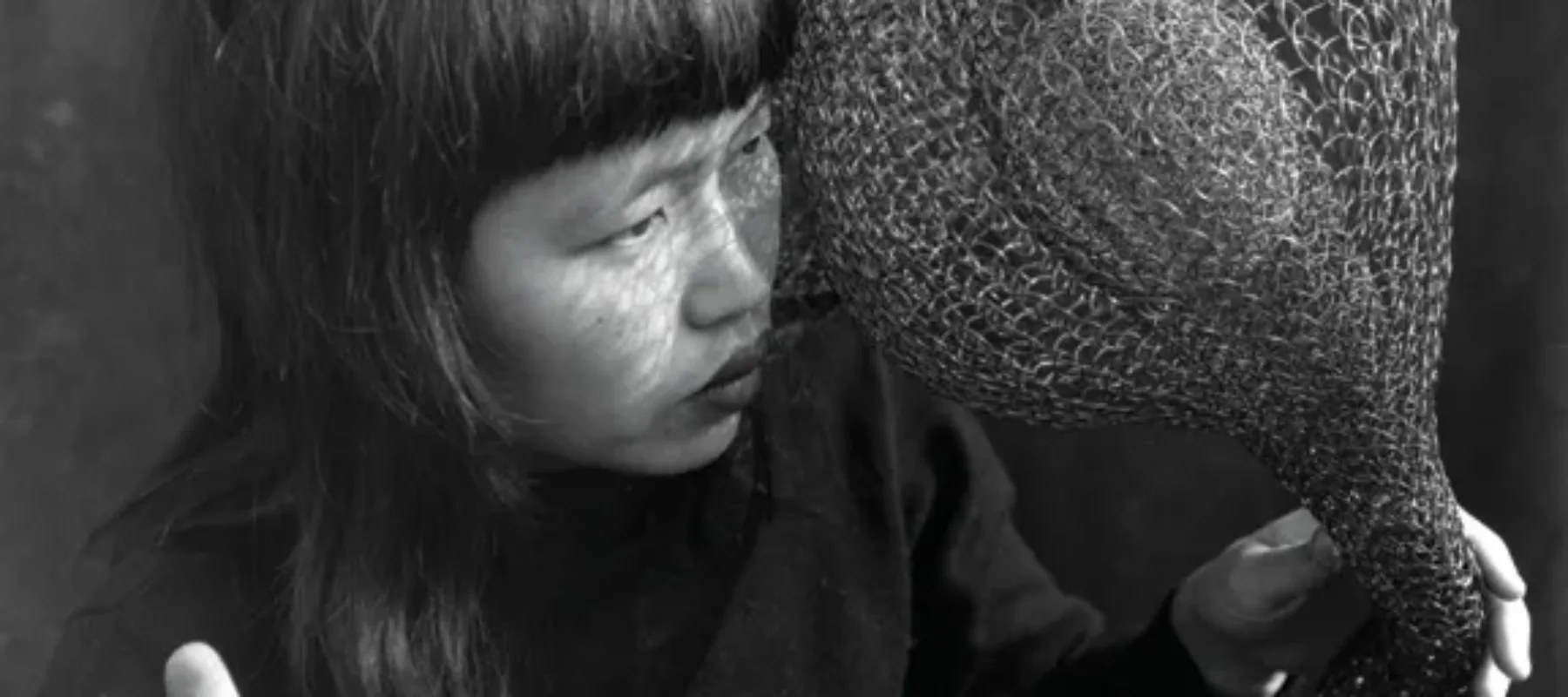 A black-and-white photograph of Ruth Asawa holding one of her large, wire crochet sculptures, draped over her shoulder and in both hands. She is a light-skinned, Asian, adult woman with black hair and blunt bangs.