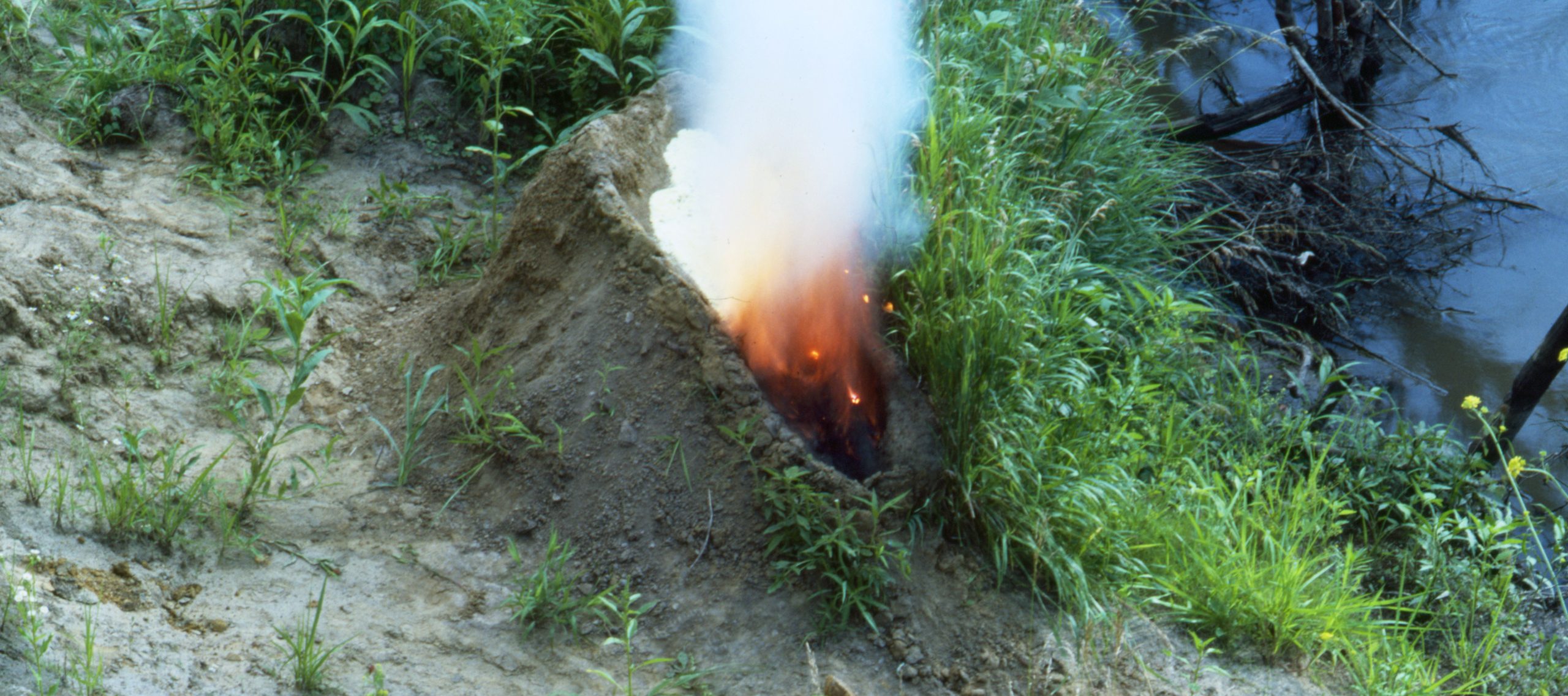 A mound of dirt in a grassy area by water. In the center of the mound is a human-shaped recess full of gunpowder that has been set aflame, exploding up in fire and white smoke, making the dirt mound resemble an erupting volcano.
