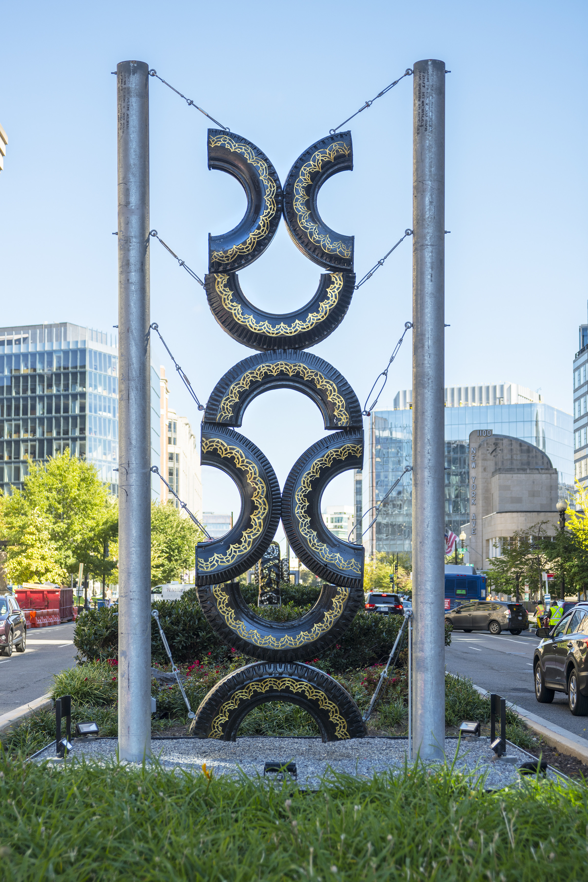 Daytime view of a city street shows a large sculpture made of eight half-tires arranged into stacked hourglass shapes. Engravings, and gold leaf decorate the structure. The sky is bright blue and cloudless.
