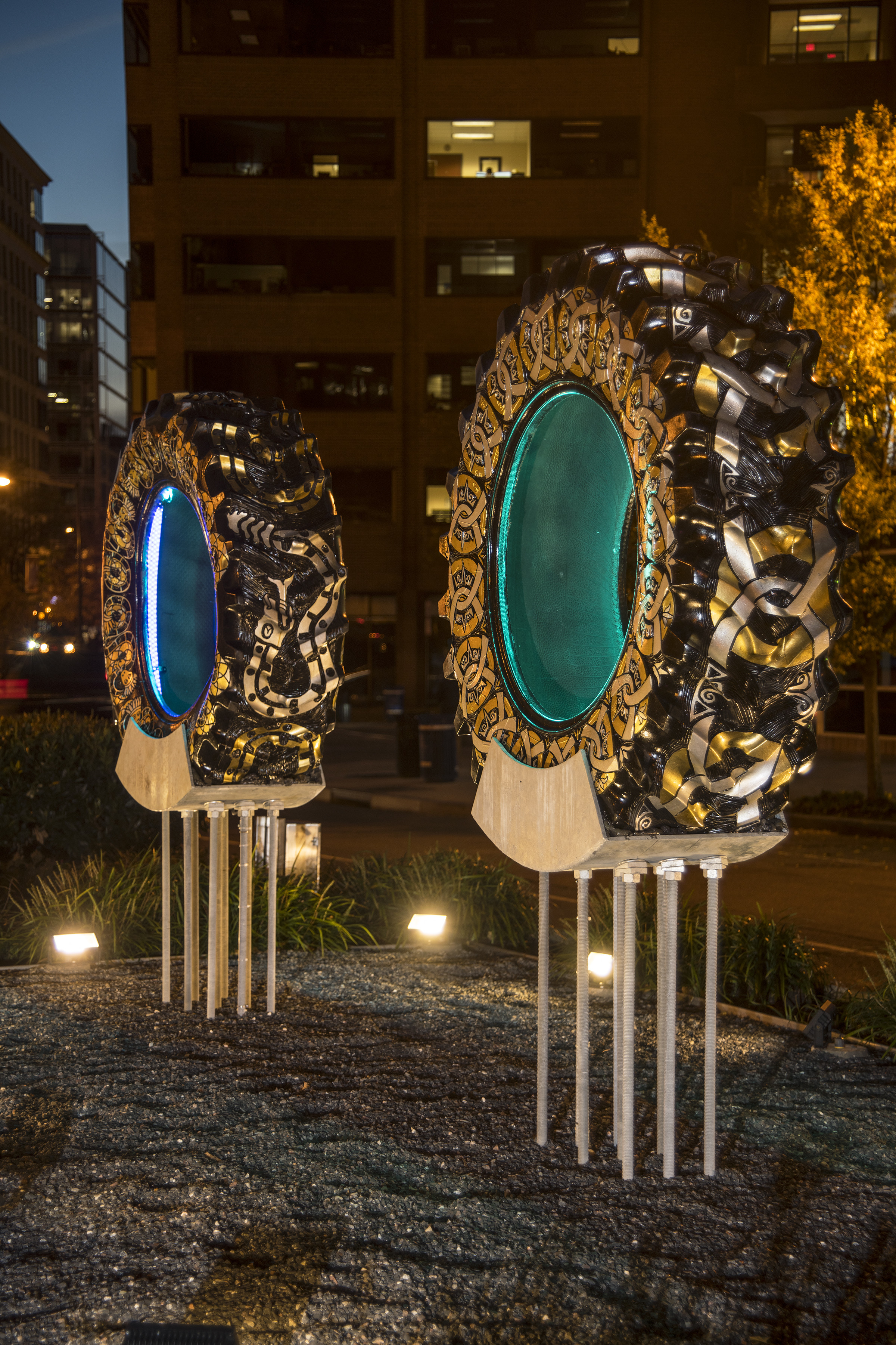 Two large tractor tires are displayed on silver legged pedestals in a center road median of a city street. Each tires is engraved with different designs: one with a snake and the other with a chain pattern. Both are designs are painted silver and gold. Blue lights placed in the interior circle of the tires light the structures up.