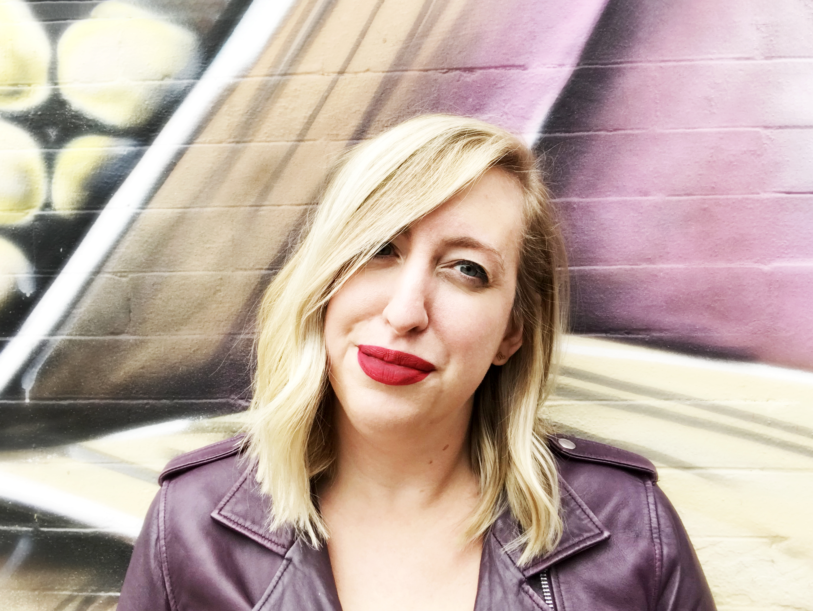 A light-skinned woman with straight shoulder-length blonde hair wears bright red lipstick and smiles at the camera. She stands in front of an abstract mural and wears a brown leather jacket.