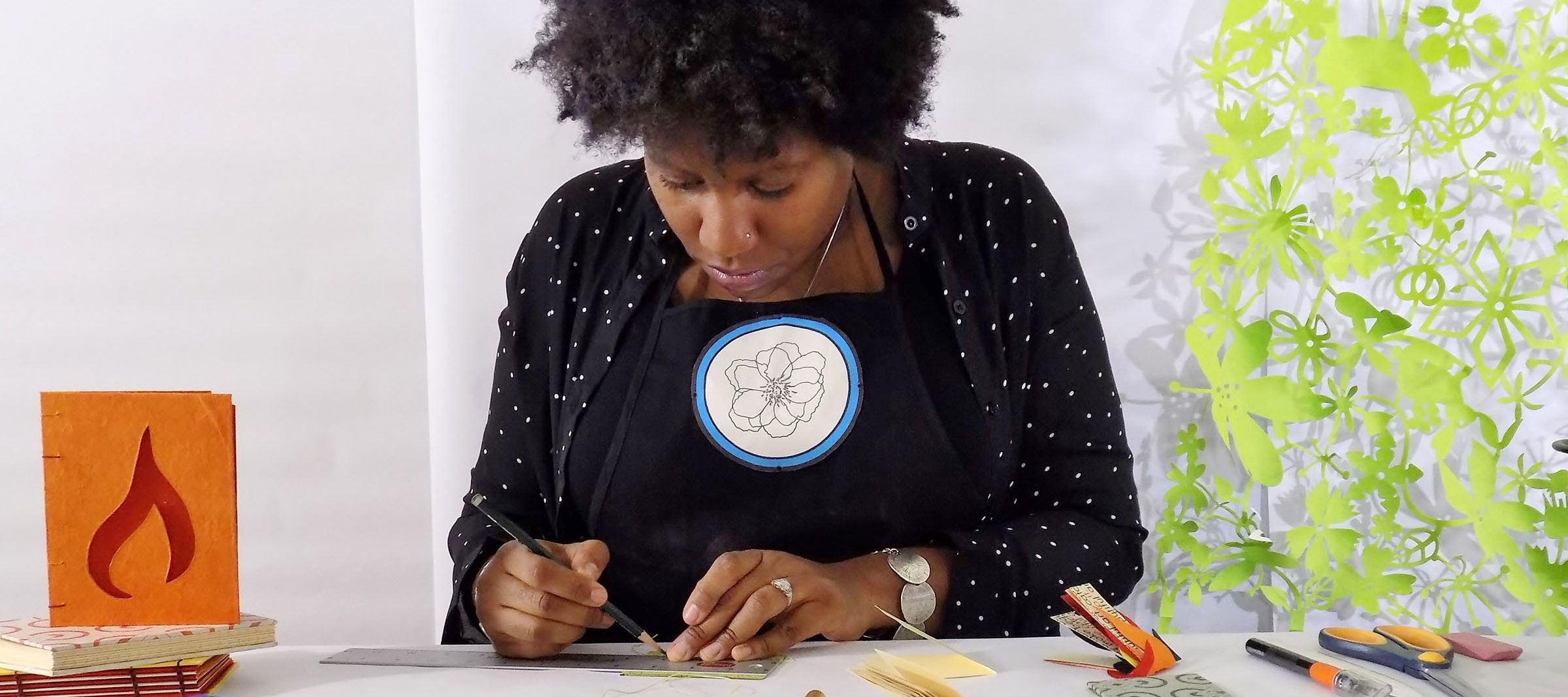 A dark-skinned adult woman holds a pencil and draws on papers on a table, with an artist's book to her right. She wears dark clothes and has a short afro.